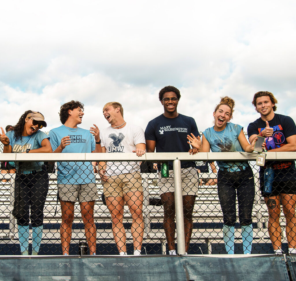 A group of students wearing University of Mary Washington Eagles T-shirts stand behind a fence, apparently at an athletics event