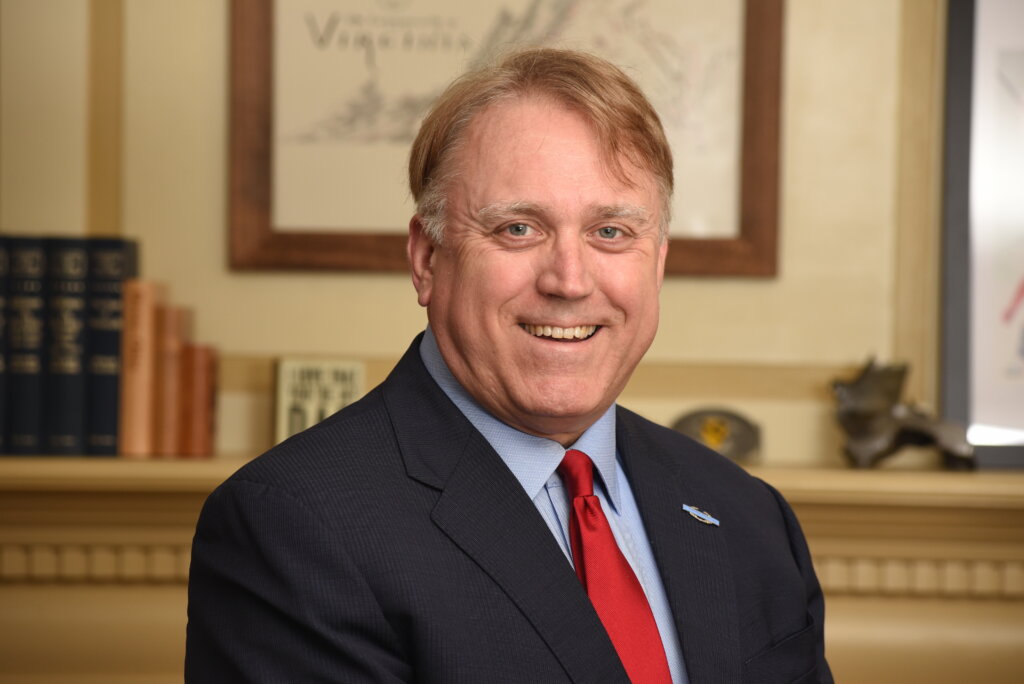 A man wearing a suit and red time smiles for the camera in front of a shelf with books
