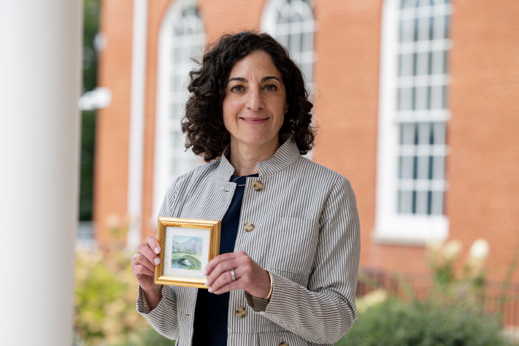 A woman stands outside of a brick building holding a small painting of a bridge