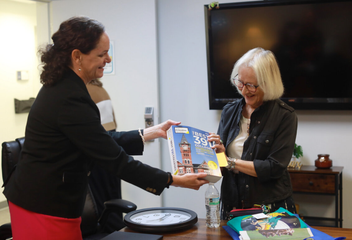 one woman with dark hair hands a book to another with light hair while standing indoors in a meeting room