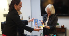 one woman with dark hair hands a book to another with light hair while standing indoors in a meeting room