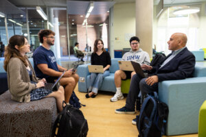 students with laptops sit in chairs next to a professor that is seated and speaking inside a building on UMW campus