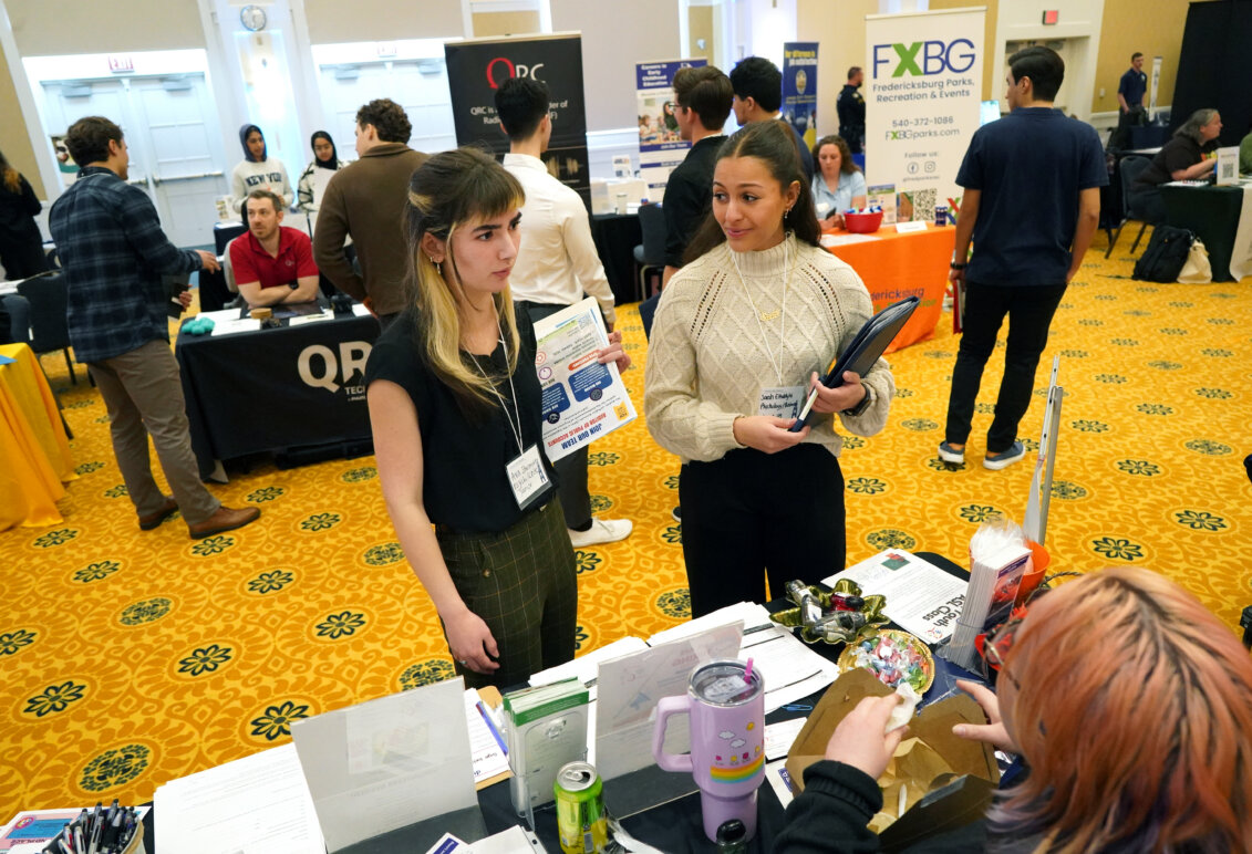 Two students, holding flyers and notebooks, stand in the middle of a busy ballroom.