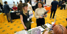 Two students, holding flyers and notebooks, stand in the middle of a busy ballroom.