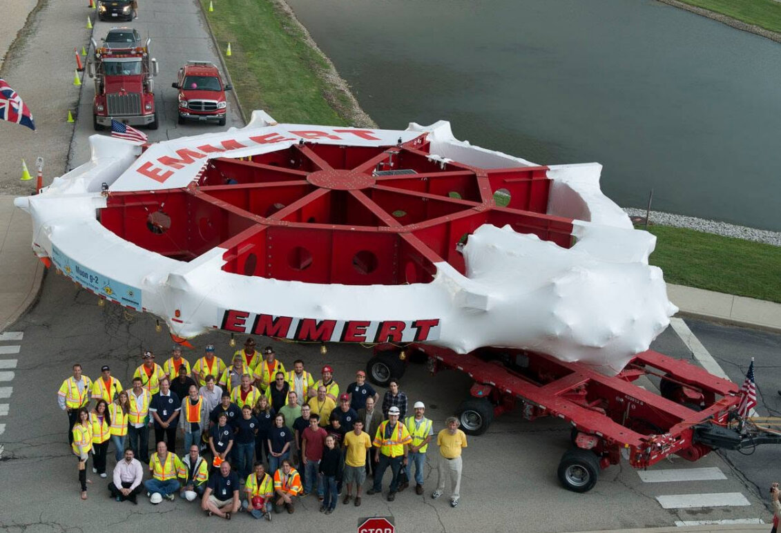A giant piece of technology on a tractor trailer, along with a large group of people, taken from an aerial view.