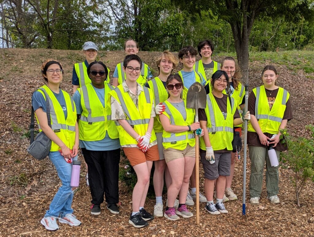 Several people stand on a leafy ground wearing neon yellow vests and holding shovels and other garden tools