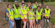 Several people stand on a leafy ground wearing neon yellow vests and holding shovels and other garden tools