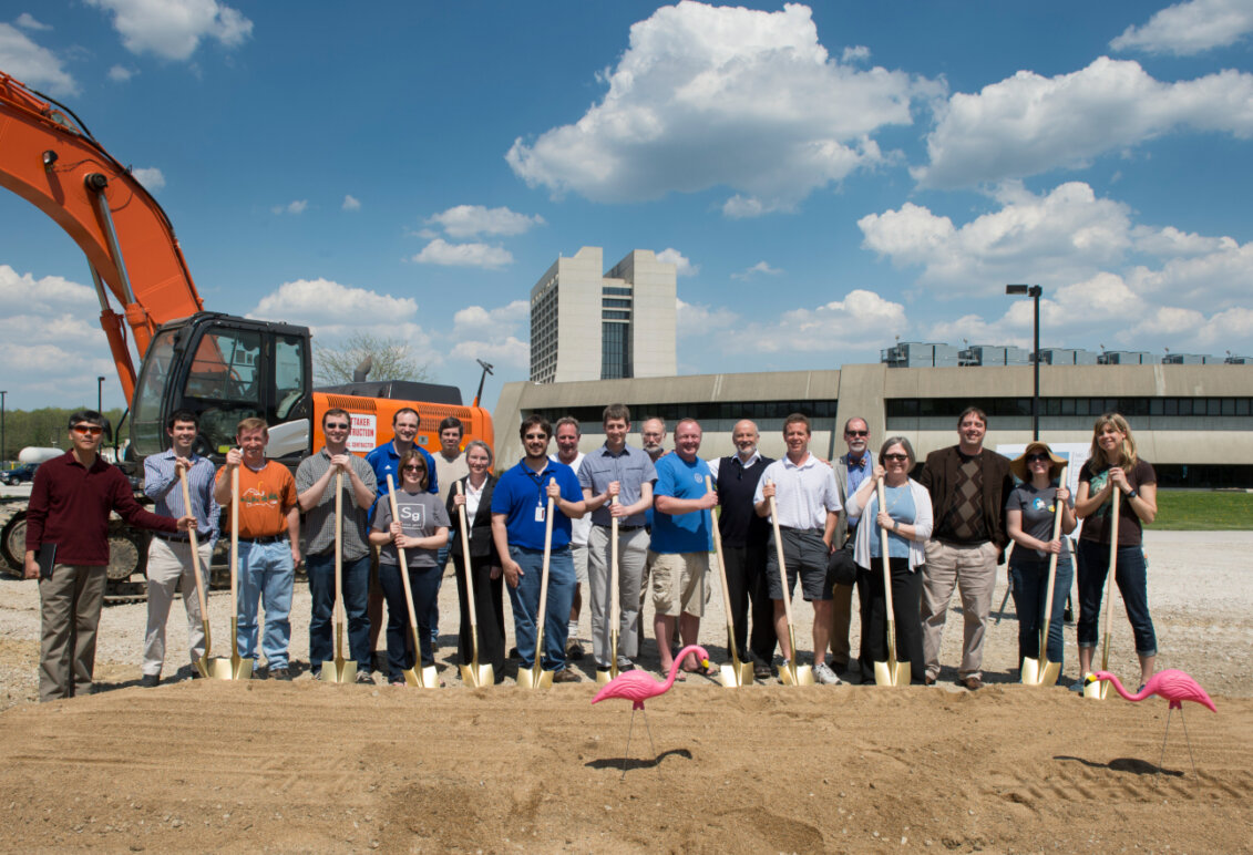 A group of people with shovels at a construction site with equipment and buildings behind them.