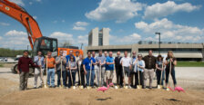 A group of people with shovels at a construction site with equipment and buildings behind them.