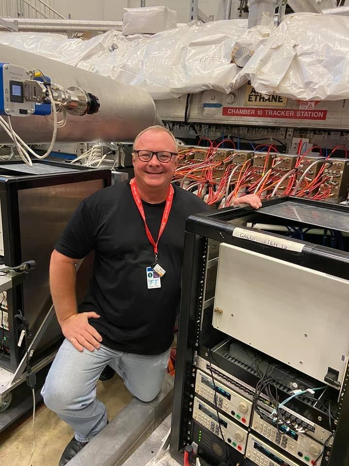 A man wearing a lanyard and smiling, surrounded by equipment for physics research.