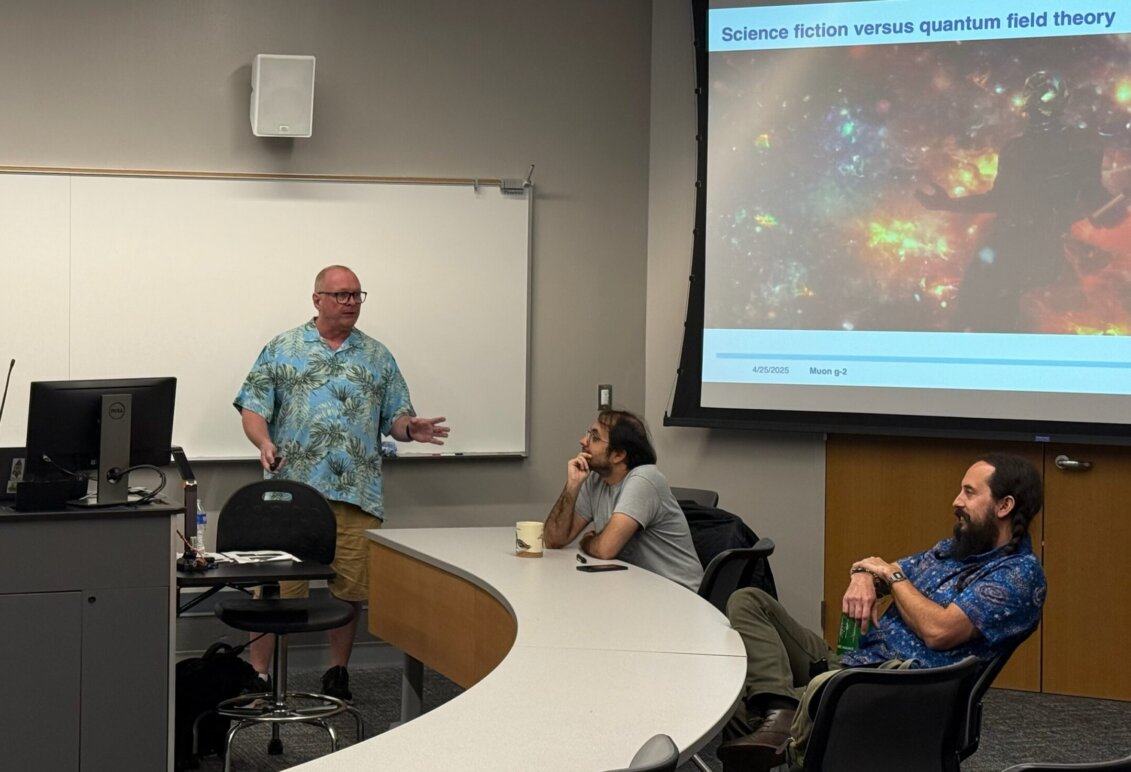 A man standing in a classroom with two professors seated at a table and a presentation on a screen.