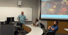 A man standing in a classroom with two professors seated at a table and a presentation on a screen.