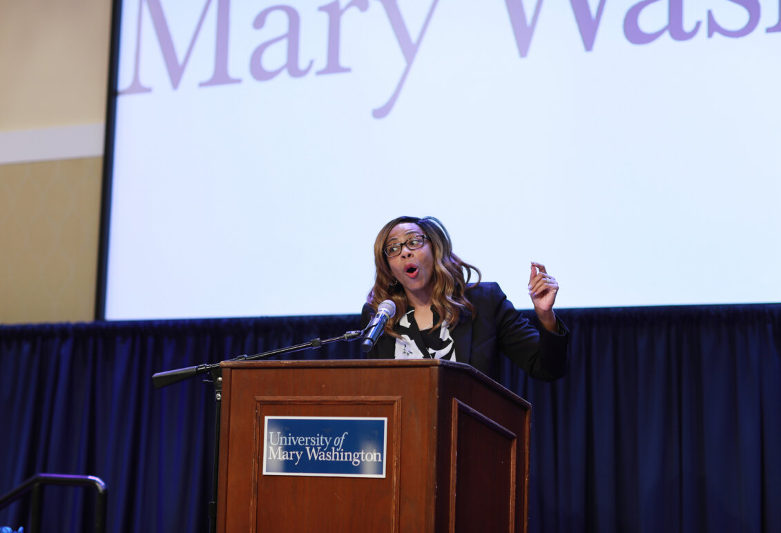 A person stands at a podium speaking into a microphone in front of a screen showing the words "University of Mary Washington."