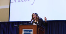 A person stands at a podium speaking into a microphone in front of a screen showing the words "University of Mary Washington."