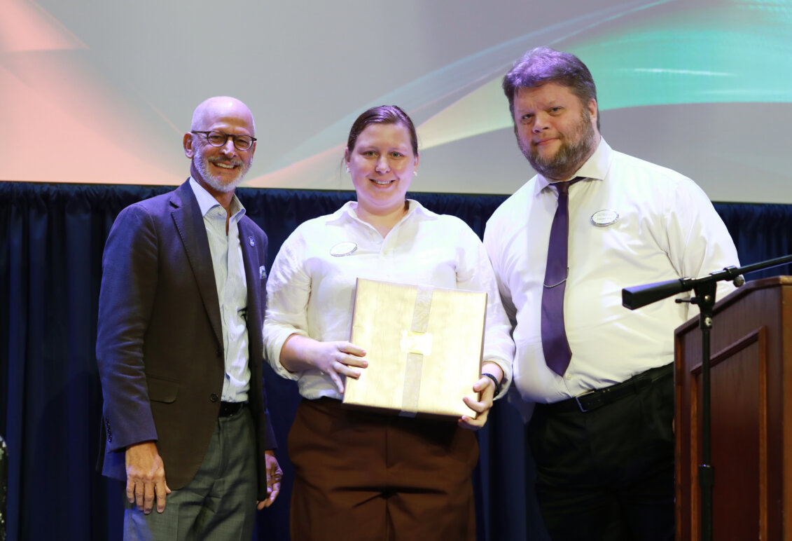 A woman stands onstage holding an award, while two men stand on either side of her