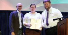 A woman stands onstage holding an award, while two men stand on either side of her