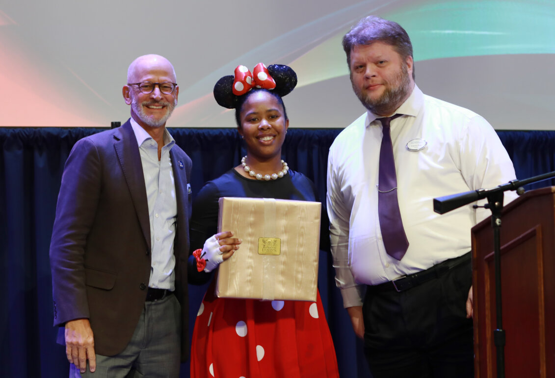 A woman in a Minnie Mouse costume stands onstage holding an award while two men stand on either side of her