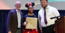 A woman in a Minnie Mouse costume stands onstage holding an award while two men stand on either side of her