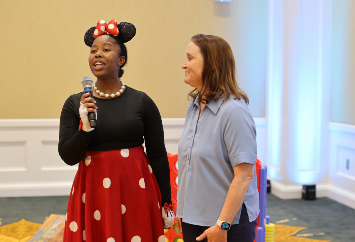 A woman dressed like Minnie Mouse and holding a microphone stands beside another woman