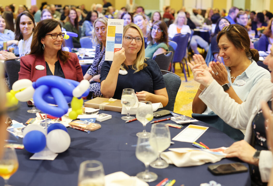 A woman holds up a prize-winning card while sitting at a table in the middle of a packed ballroom.