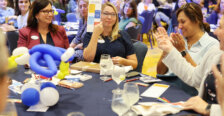 A woman holds up a prize-winning card while sitting at a table in the middle of a packed ballroom.