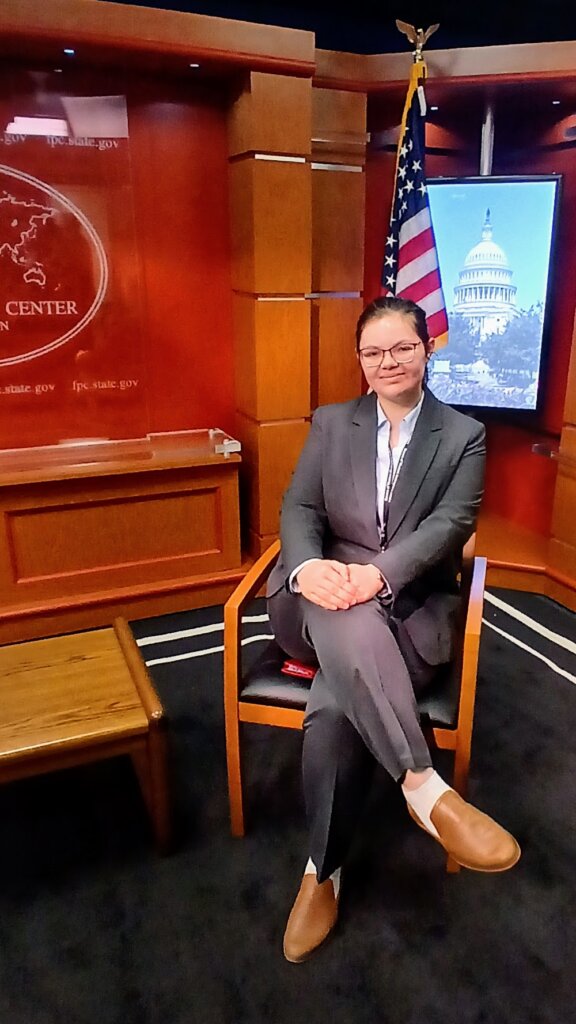 A UMW student in a suit and glasses, seated in a chair in the U.S. Department of State with a red background and an American flag behind her.