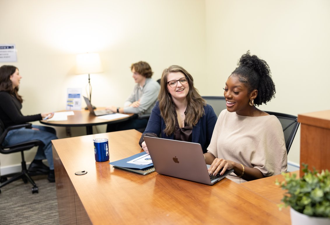 Two people sit behind a desk looking at a laptop in the foreground while two students sit at a table across from each other in the background