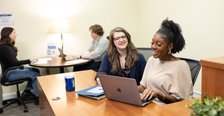Two people sit behind a desk looking at a laptop in the foreground while two students sit at a table across from each other in the background
