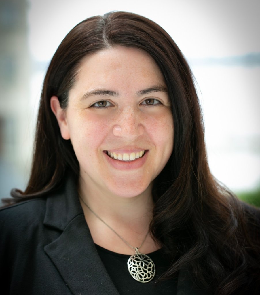 A photo of a smiling person with long, dark brown hair and a black suit jacket and shirt with a silver medallion necklace.