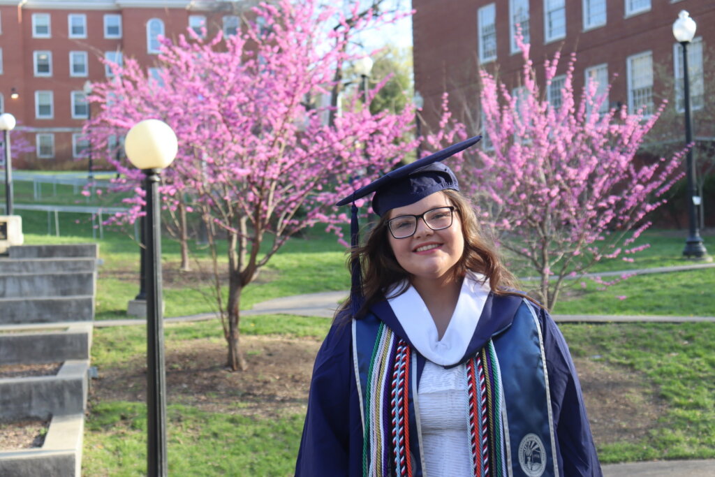 A student in a blue cap and gown, smiling and standing on the UMW campus with brick buildings and pink blossoms in the background.