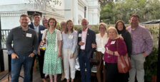 A group of 11 smiling people on a deck, with several holding wine glasses.