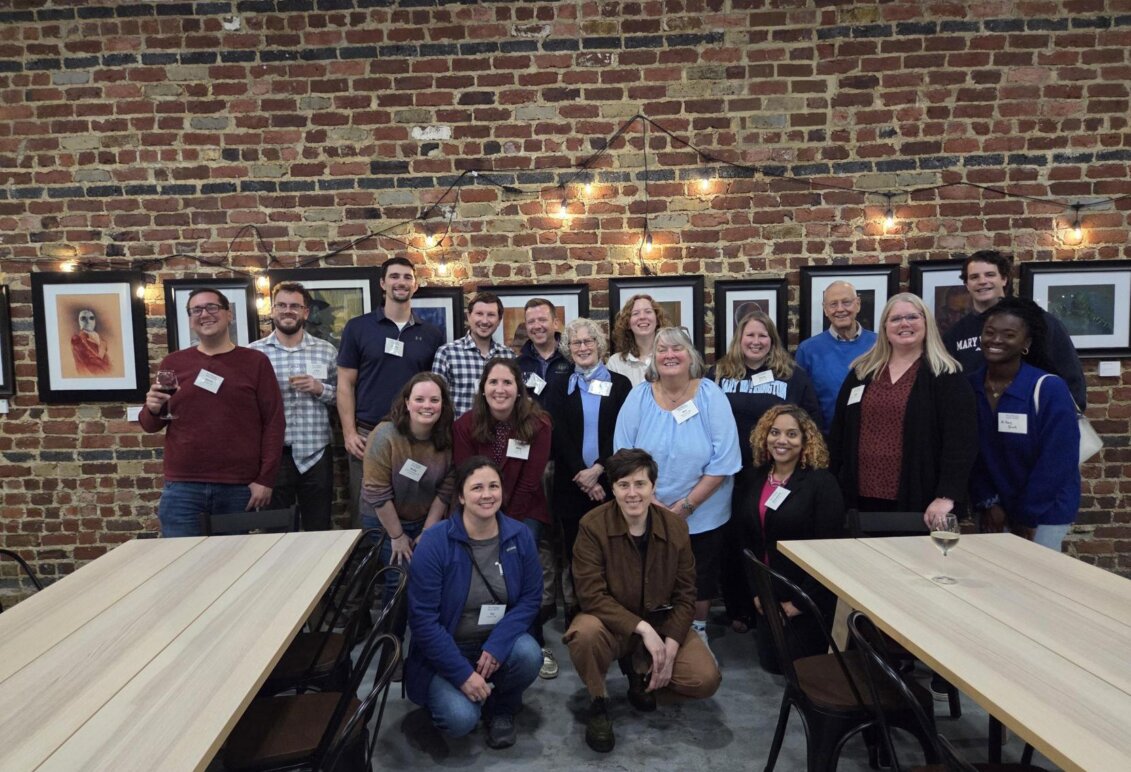 A large group of people standing against a brick wall in a winery. Paintings are on the wall, lights are overhead, and two tables flank the group.