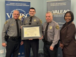 three male police officers in uniforms and a woman in brown suit stand together holding a certificate of accreditation