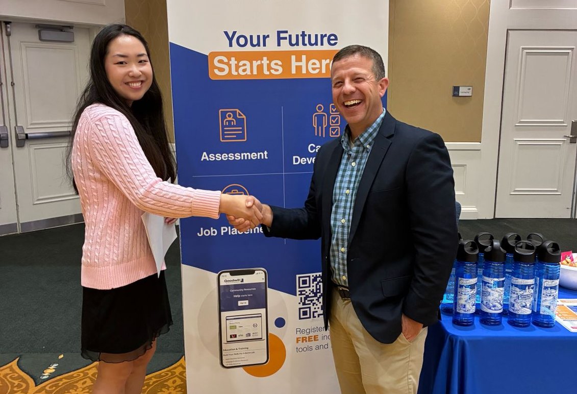 A man and a female student shake hands in front of a sign for Goodwill Industries at a Career Fair tabling event.