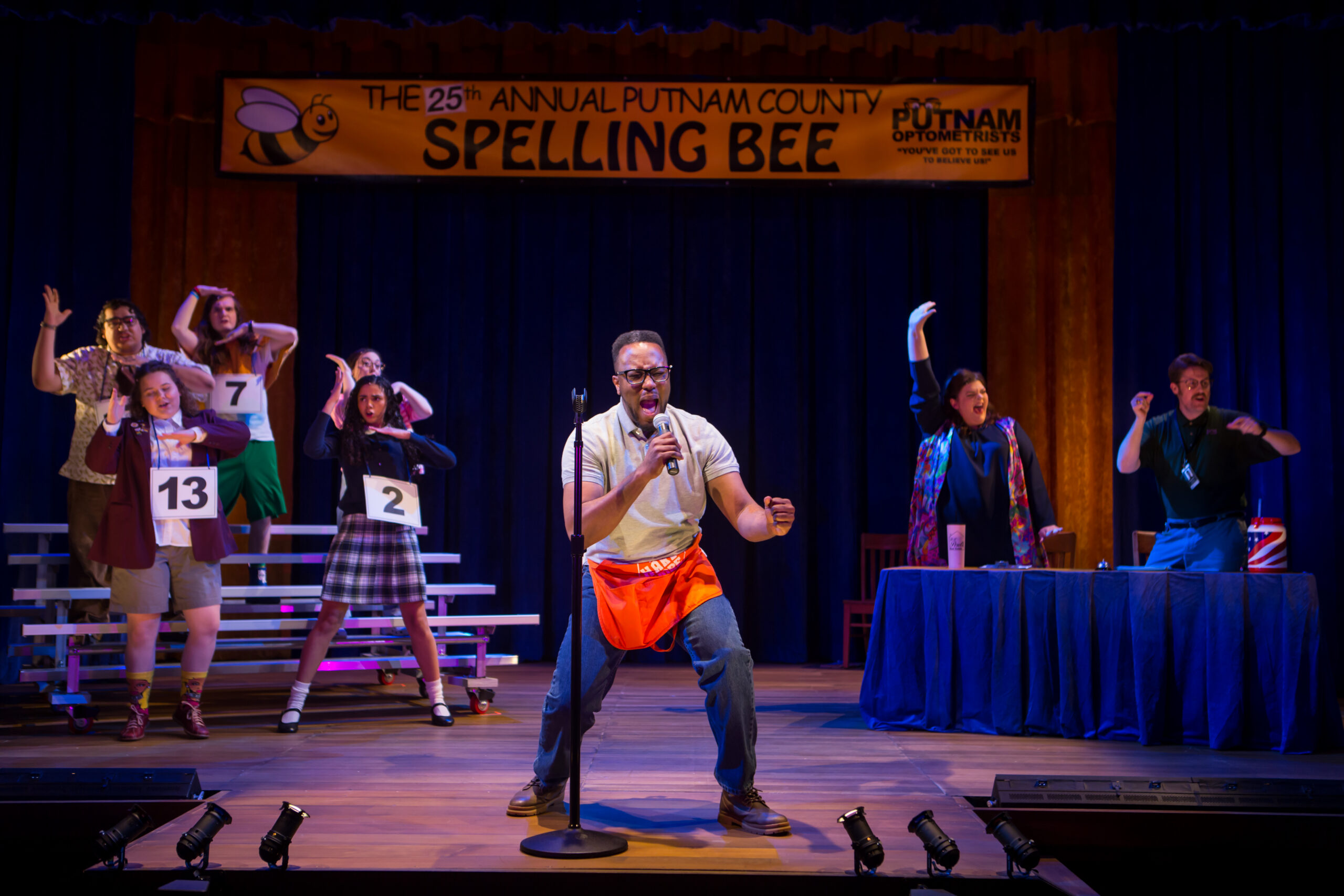 Actos performing on stage in "The 25th Annual Putnam County Spelling Bee". A soloist is downstage center holding a microphone and singing.