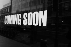 
A black-and-white, low-angle photo of a shop window with the words "COMING SOON" in large, bold, white letters reflected on the glass. The reflection is slightly distorted and blurry, with the street and buildings behind the viewer visible.